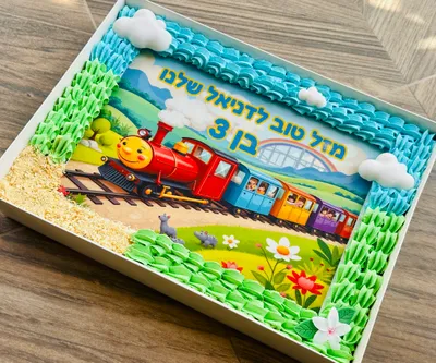 עוגת גן רכבת מלבנית, עם דף סוכר של רכבת צבעונית, מוקפת זילופי קרם ירוקים ותכולים, מלמעלה ובזווית קלה